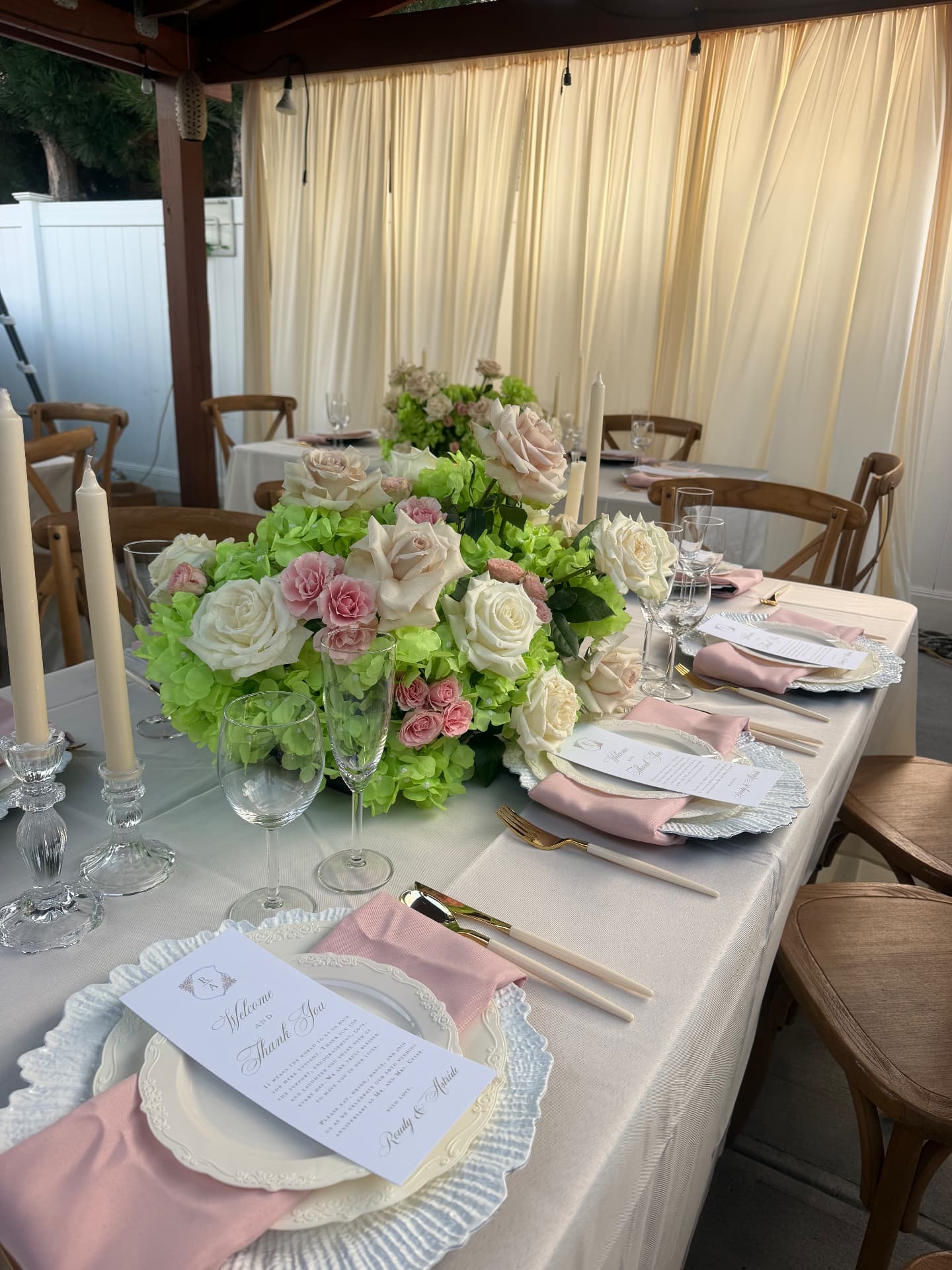 Outdoor event tablescape with ivory and blush rose centerpiece, gold flatware and draped backdrop